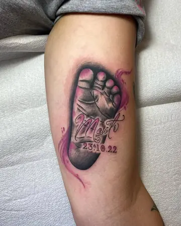 Tatuagem realista de um pezinho de bebê com nome em script e a data 23.10.22, com detalhe rosa