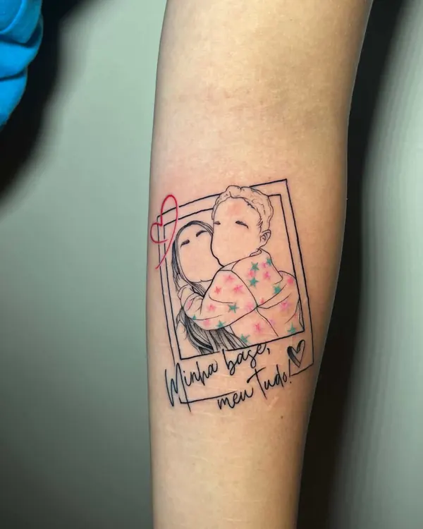 Antebraço com tatuagem fine line de mãe e filho abraçados numa moldura de polaroid e a frase Minha base, meu tudo
